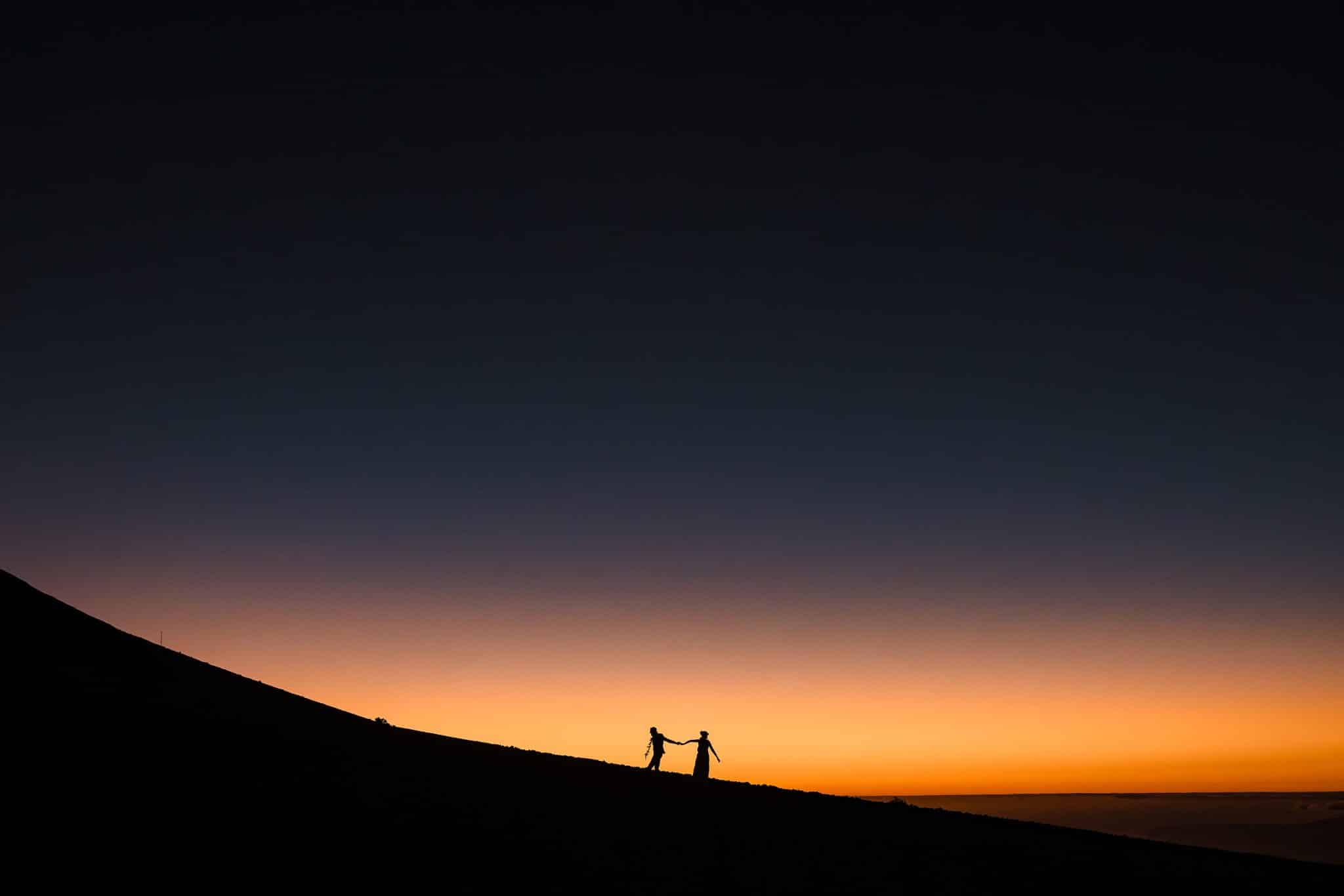 Cheryl and Lincoln's sillohouette walking among Haleakala as the sky is orange and black during the sunset