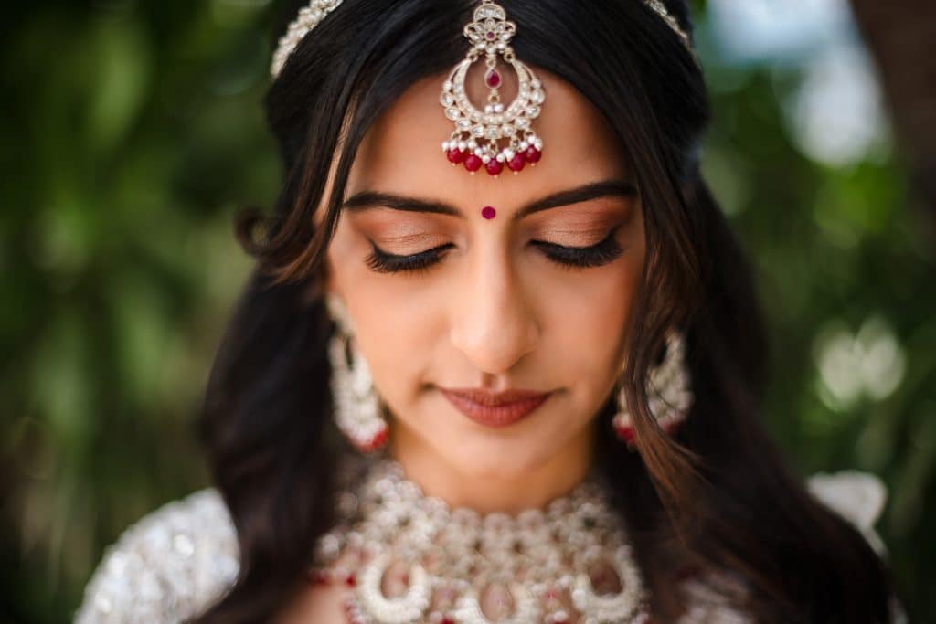 Indian-Wedding-in-Maui-_0005 - Ann Arbor Wedding Photographer Close-up of a woman with intricate silver jewelry and red gems, eyes closed, wearing a decorative forehead piece (maang tikka).