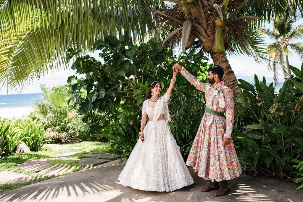 Indian-Wedding-in-Maui-_0009 - Ann Arbor Wedding Photographer A photo of a groom spinning his bride in her wedding attire while they stand under a palm tree. Surrounded by greenery and the ocean peeking from behind.
