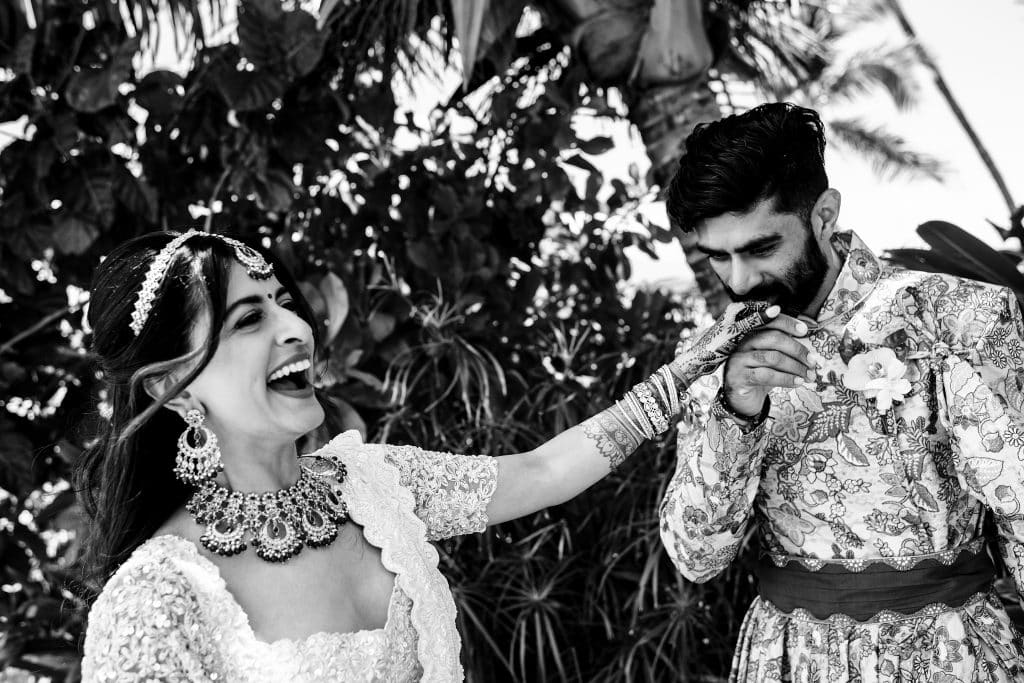 Indian-Wedding-in-Maui-_0010 - Ann Arbor Wedding Photographer Joyful bride in ornate jewelry laughing as the groom adjusts his embroidered sherwani outdoors.