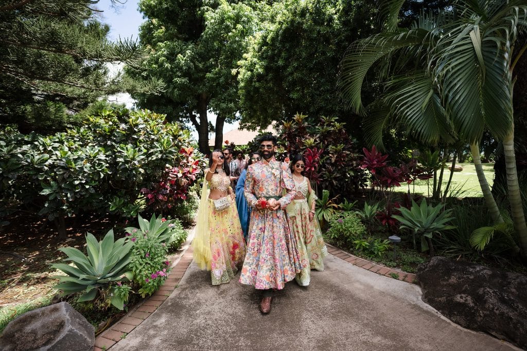 Indian-Wedding-in-Maui-_0011 - Ann Arbor Wedding Photographer A groom in his embroidered sherwani as wedding guests trail behind him on a path lined with lush greenery.