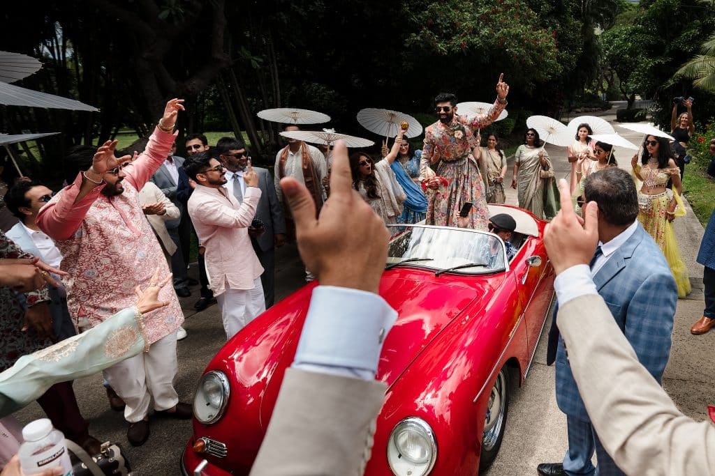 Indian-Wedding-in-Maui-_0012 - Ann Arbor Wedding Photographer Wedding guests celebrate around a bright red vintage convertible, some holding white parasols and raising hands in joy.