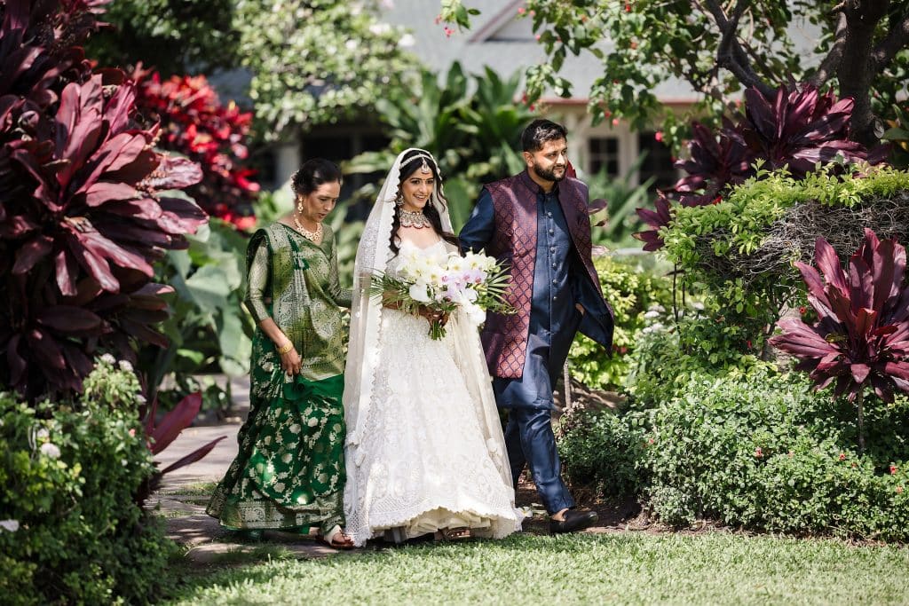 Indian-Wedding-in-Maui-_0016 - Ann Arbor Wedding Photographer Bride in a white wedding gown holding a bouquet walks beside a groom in a blue and maroon outfit, with a woman in green beside them in a lush garden.