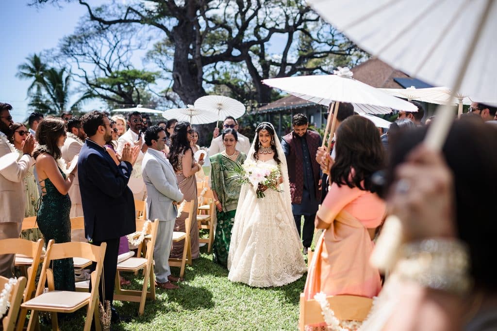 Indian-Wedding-in-Maui-_0017 - Ann Arbor Wedding Photographer Bride in a white wedding gown holding a bouquet walks beside a groom in a blue and maroon outfit, with a woman in green beside her as they walk down the aisle. Guests are standing and holding parasols as they watch.