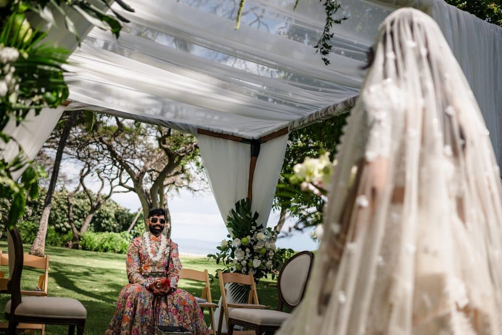 Indian-Wedding-in-Maui-_0018 - Ann Arbor Wedding Photographer Outdoor wedding setup with white draped fabric and floral arch under trees; a man in a colorful floral suit sits among wooden chairs.