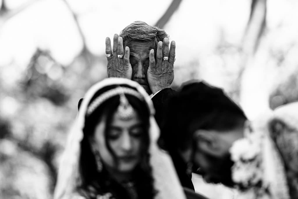Indian-Wedding-in-Maui-_0021 - Ann Arbor Wedding Photographer A man holding his face in both hands as he wipes away tears is in focus. A bride and groom looking down at the ground is in the foreground out of focus.