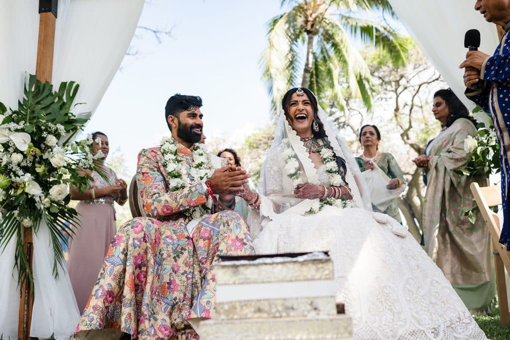 Indian-Wedding-in-Maui-_0022 - Ann Arbor Wedding Photographer Bride and groom laughing on a stone staircase during an outdoor wedding, surrounded by family in floral attire and white flowers.