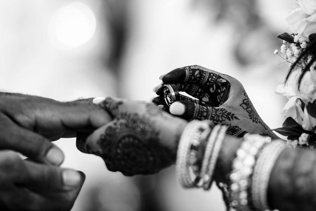Indian-Wedding-in-Maui-_0023 - Ann Arbor Wedding Photographer Close-up of a henna-decorated hand placing a ring on another hand during a wedding ceremony, with a floral garland in the frame.