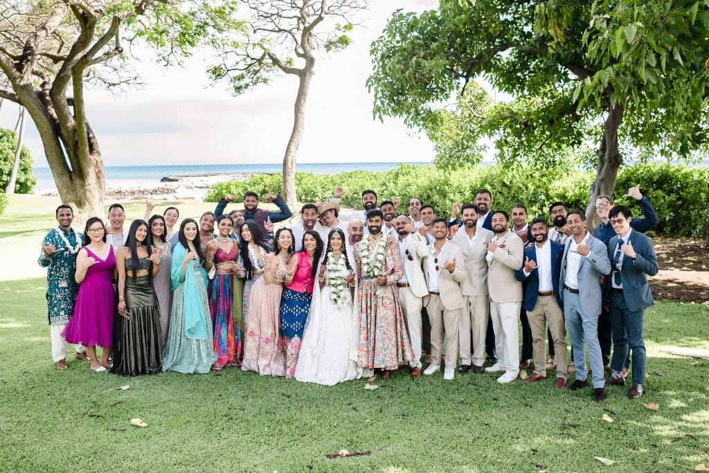 Indian-Wedding-in-Maui-_0024 - Ann Arbor Wedding Photographer A group photo of a bride and groom adorned in white florals as the wedding guests surround them and smile at the camera with the beach not far behind them.