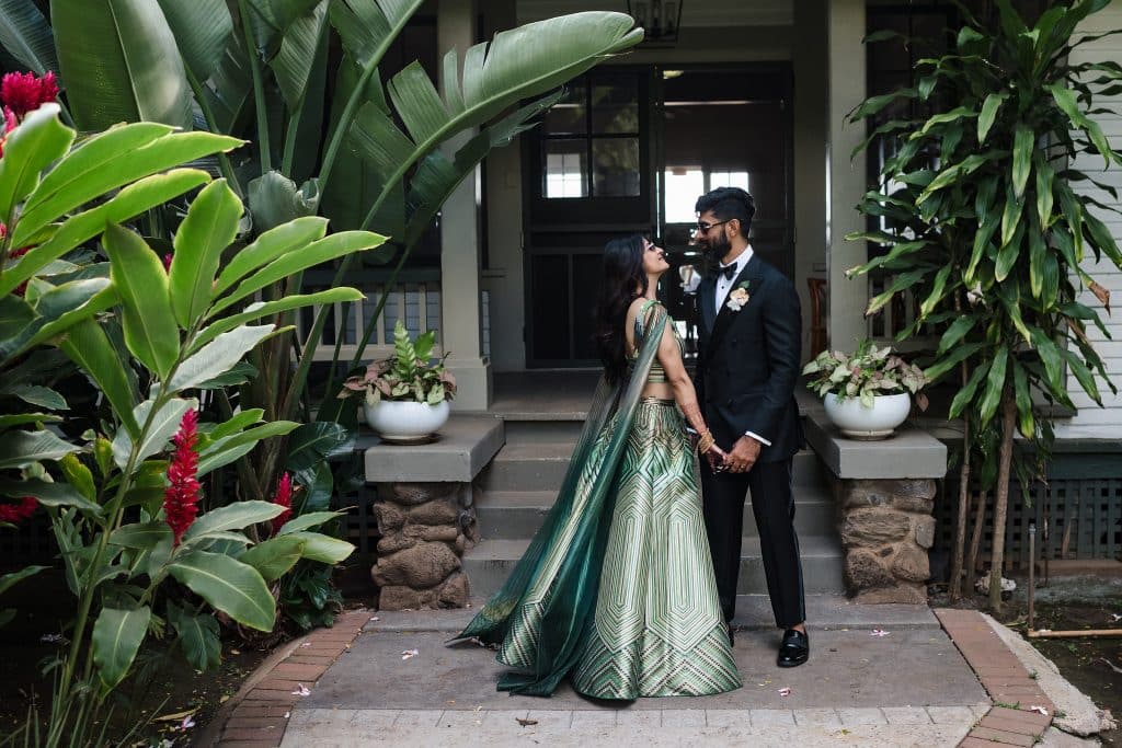 Indian-Wedding-in-Maui-_0025 - Ann Arbor Wedding Photographer A couple stands on a front staircase surrounded by tropical plants; the woman wears a green patterned saree, the man in a black suit and bow tie, holding hands and gazing at each other.