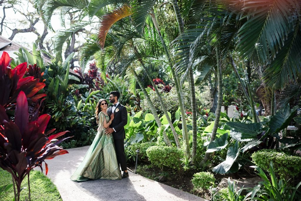 Indian-Wedding-in-Maui-_0026 - Ann Arbor Wedding Photographer A couple dressed in formal attire stands together on a garden path among tropical plants and red-leaf foliage.
