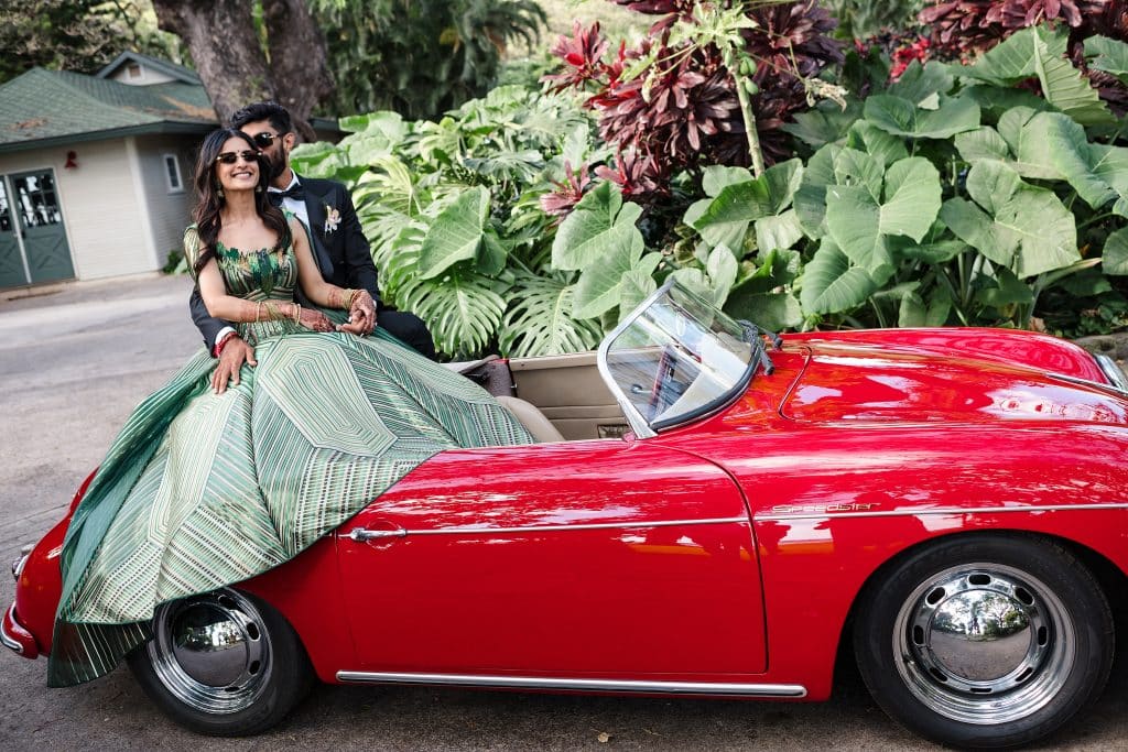 Indian-Wedding-in-Maui-_0027 - Ann Arbor Wedding Photographer A couple sits in a bright red vintage convertible: woman in a green patterned gown, man in a dark suit wearing sunglasses, tropical plants in the background.