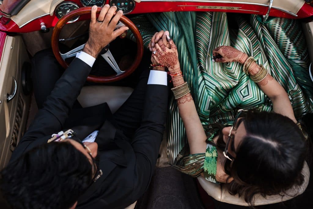 Indian-Wedding-in-Maui-_0028 - Ann Arbor Wedding Photographer A birds-eye view of a riding in a red vintage convertible car, holding hands. The woman is wearing a green dress with henna designs on her hands. The man is wearing a black suit.