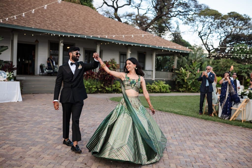 Indian-Wedding-in-Maui-_0032 - Ann Arbor Wedding Photographer A groom spinning his bride who is wearing a green top and skirt on a brick patio with string lights above. Wedding guests are clapping behind them as they enter the reception.