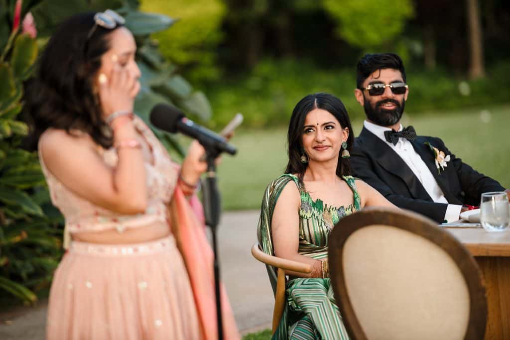 Indian-Wedding-in-Maui-_0035 - Ann Arbor Wedding Photographer Couple sitting at a table and smiling at a wedding guest wearing peach who is making a toast into a microphone.