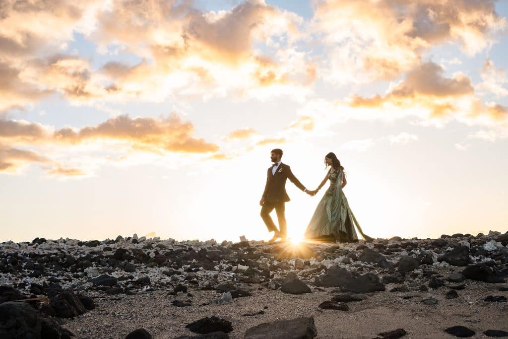 Indian-Wedding-in-Maui-_0039 - Ann Arbor Wedding Photographer A couple in formal Indian attire holding hands while walking on a rocky beach at sunset