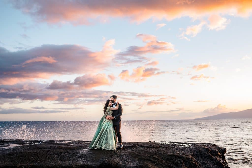 Indian-Wedding-in-Maui-_0041 - Ann Arbor Wedding Photographer Couple hugging on a rocky shoreline of the ocean while the sun sets and waves splash behind them.