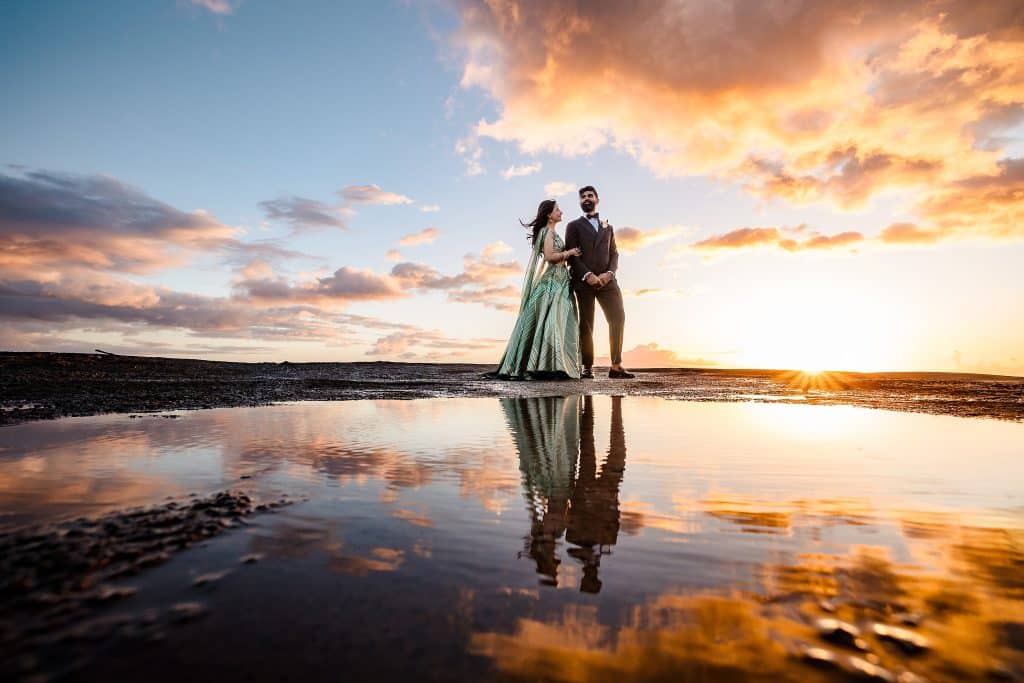 Indian-Wedding-in-Maui-_0042 - Ann Arbor Wedding Photographer A formally dressed couple stands on a beach at sunset, the woman in a green gown and the man in a dark suit, holding hands with a reflective pool at their feet.
