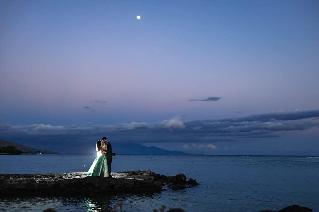 Indian-Wedding-in-Maui-_0043 - Ann Arbor Wedding Photographer Couple dancing on a rocky shoreline at dusk, moon in a blue twilight sky above a calm sea.