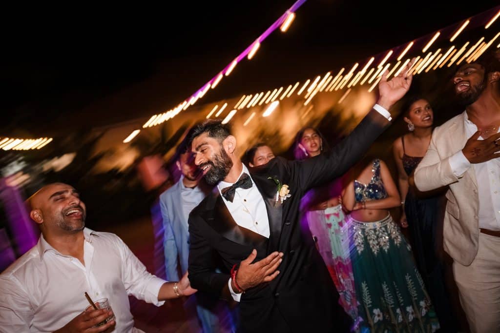 Indian-Wedding-in-Maui-_0046 - Ann Arbor Wedding Photographer Group of friends dancing at night under warm string lights, wearing formal and festive attire with joyful expressions and motion blur.