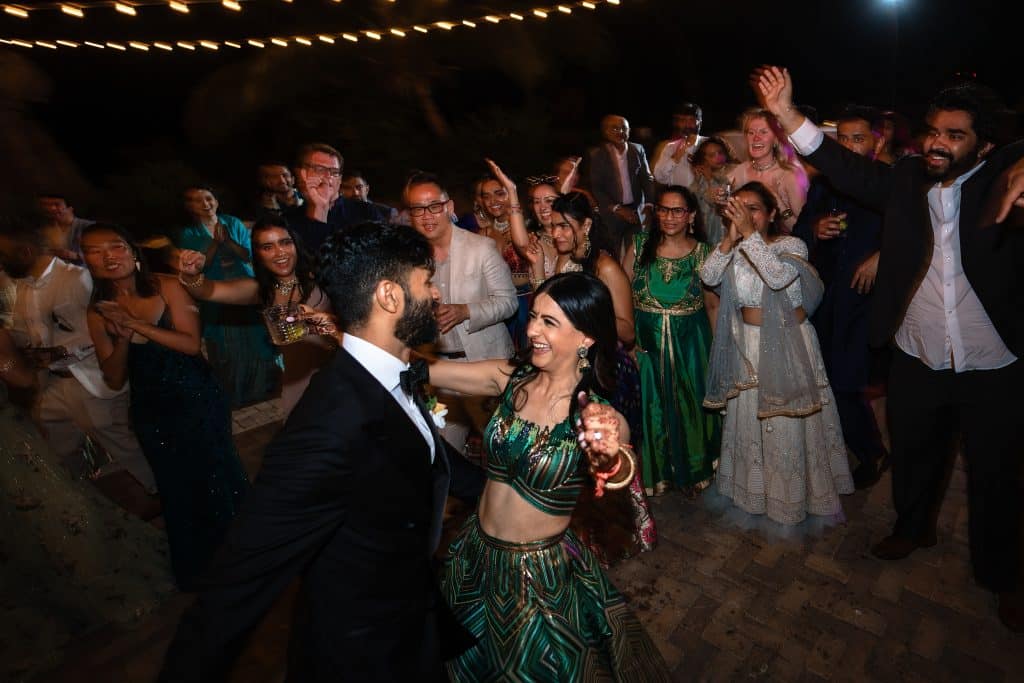 Indian-Wedding-in-Maui-_0047 - Ann Arbor Wedding Photographer Couple dressed in formal Indian attire dancing and laughing at an outdoor wedding reception lined with lights. Wedding guests are clapping and cheering behind them.