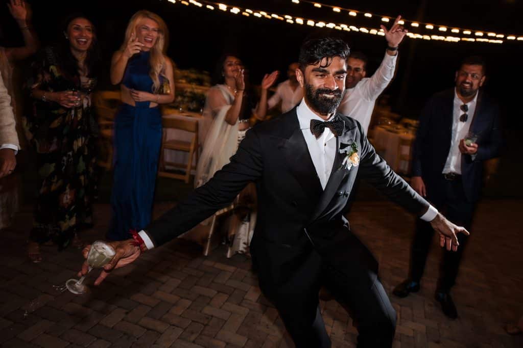 Indian-Wedding-in-Maui-_0048 - Ann Arbor Wedding Photographer Man in a black tuxedo dancing with a wine glass in his hand at an outdoor evening party, string lights overhead and cheering guests behind him.