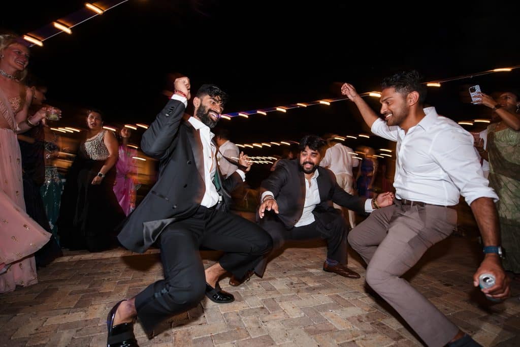 Indian-Wedding-in-Maui-_0050 - Ann Arbor Wedding Photographer Two men in a black suit and another man in formal attire are dancing low to the ground with their hands in the air.