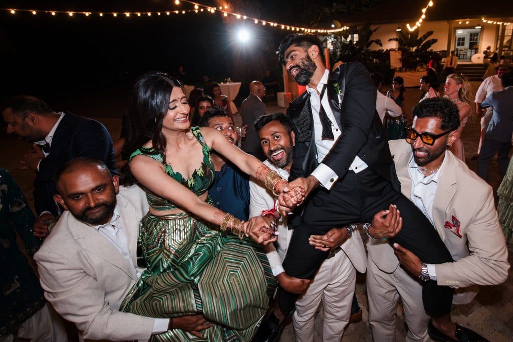 Indian-Wedding-in-Maui-_0051 - Ann Arbor Wedding Photographer Group of friends dancing joyfully at an outdoor night reception under string lights, wearing formal attire and bright dresses.